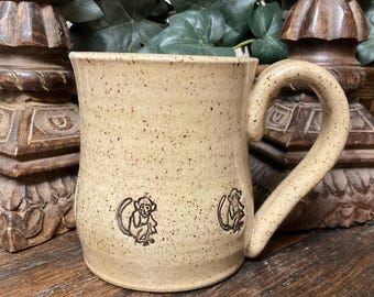 May include: A beige ceramic mug with a speckled finish and a curved handle. The exterior surface features two small, dark brown stamped illustrations of monkeys positioned near the base of the mug, evenly spaced around the cylindrical body.