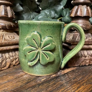 May include: A green ceramic mug with a speckled finish and a raised, embossed four-leaf clover design on the front. The mug features a sturdy, curved handle and a smooth, glossy glaze that highlights the textured details of the shamrock motif.