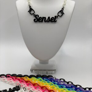 May include: A black plastic necklace with the word "Senset" in a cursive font. The necklace is on a white mannequin. Below the mannequin are multiple chains in various colors, including black, white, pink, red, orange, yellow, green, blue, purple, and light blue.