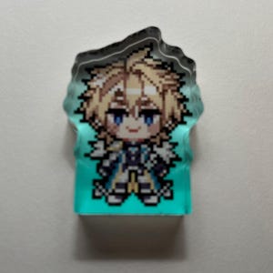 May include: A pixelated character design on a teal background. The character has blonde hair, blue eyes, and wears a blue and white outfit. The top edge is a dark, irregular shape, possibly a keychain or charm.