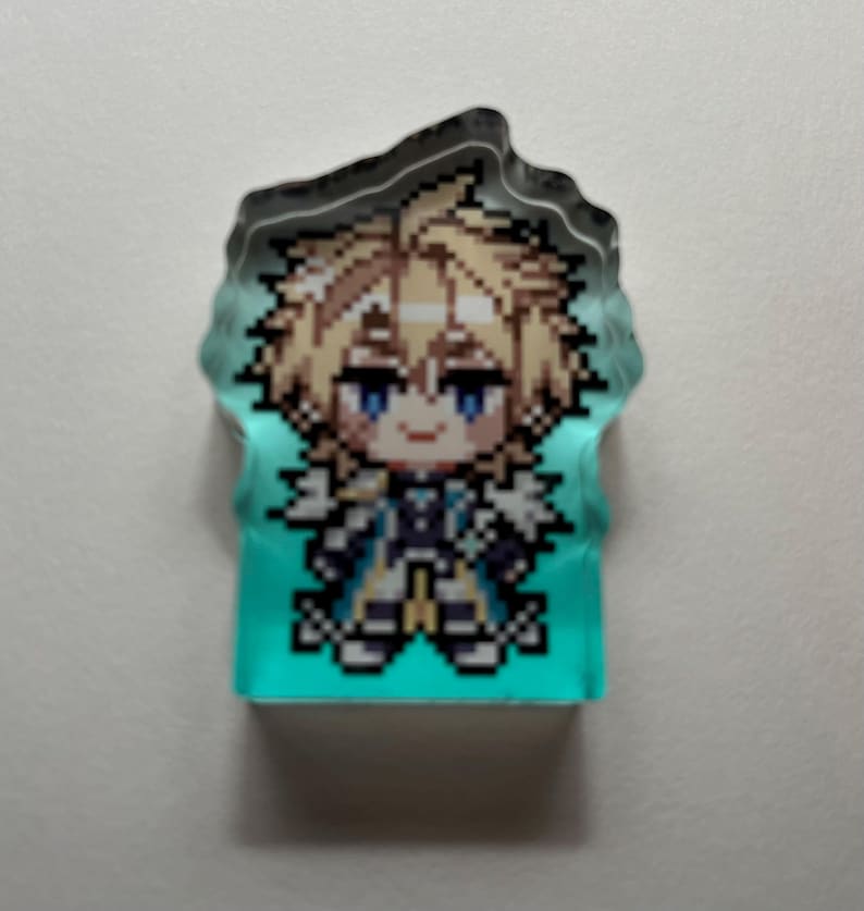 May include: A pixelated character design on a teal background. The character has blonde hair, blue eyes, and wears a blue and white outfit. The top edge is a dark, irregular shape, possibly a keychain or charm.