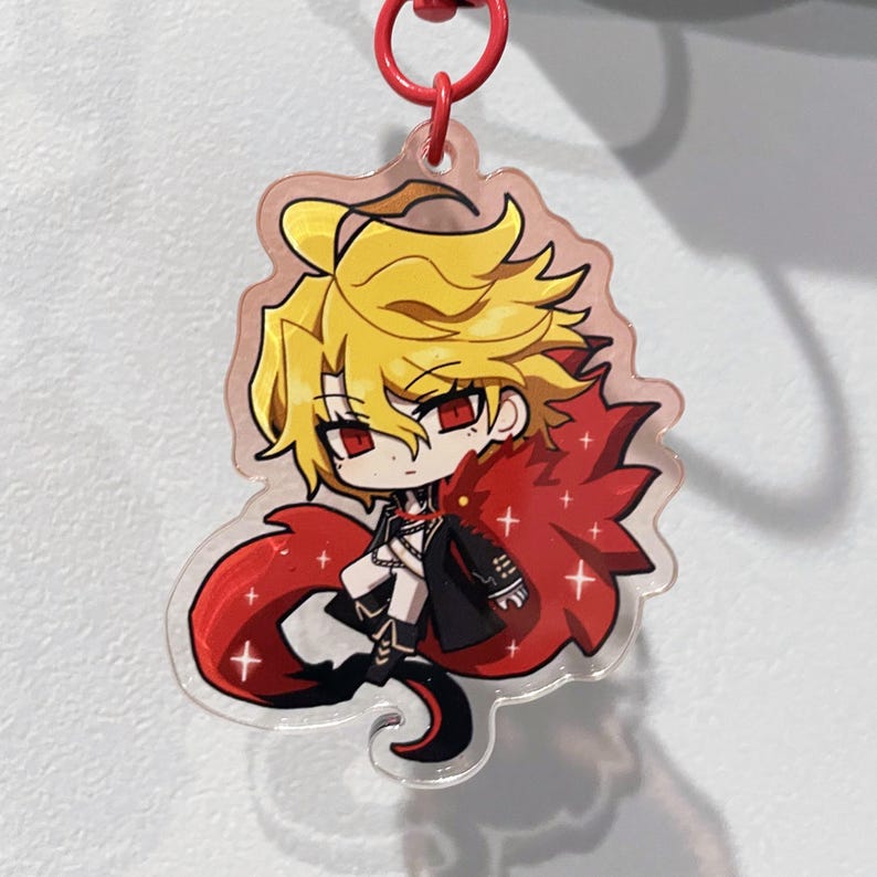May include: A clear acrylic keychain featuring a cartoon character with yellow hair, red eyes, and a red cape. The character is wearing a black outfit and has a red keychain ring attached.