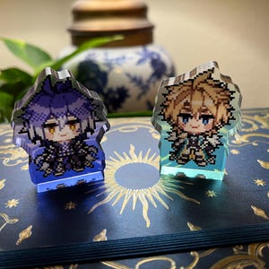May include: Two pixelated character figurines, one with purple hair and the other with blonde hair, are displayed. They are illuminated with blue and green light, respectively. The figurines are on a dark blue surface with gold accents.