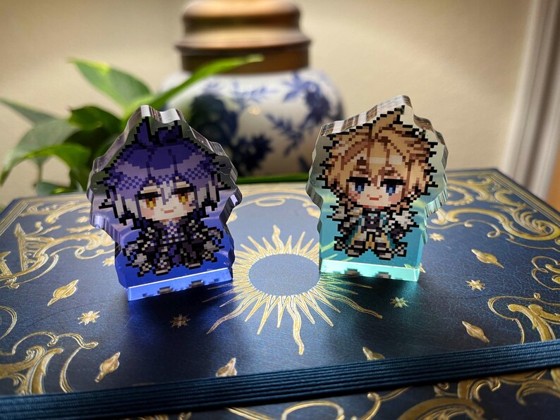 May include: Two pixelated character figurines, one with purple hair and the other with blonde hair, are displayed. They are illuminated with blue and green light, respectively. The figurines are on a dark blue surface with gold accents.