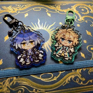 May include: Two acrylic keychains featuring pixelated anime-style characters. One keychain has a blue and black color scheme, while the other is green and gold. Both have a decorative border and a metal clasp.