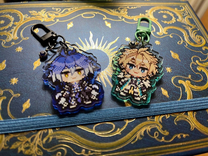 May include: Two acrylic keychains featuring pixelated anime-style characters. One keychain has a blue and black color scheme, while the other is green and gold. Both have a decorative border and a metal clasp.