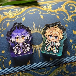 May include: Two pixel art style acrylic charms. One charm features a character with purple hair and clothing, the other has blonde hair and teal clothing. Both charms are on a blue background with gold embellishments.