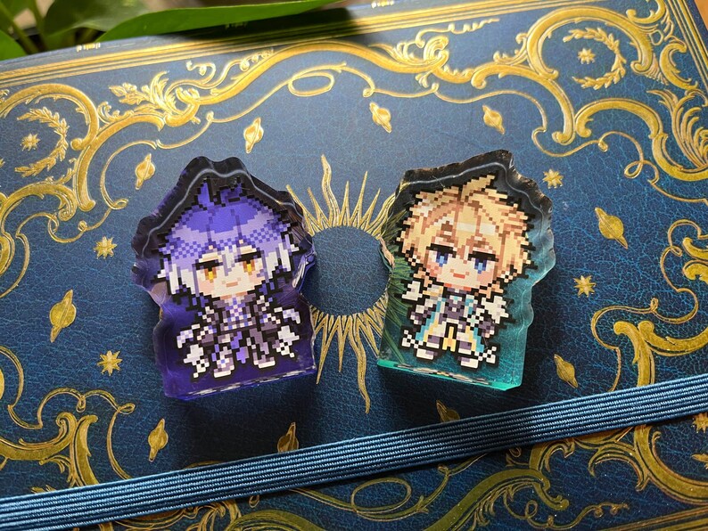 May include: Two pixel art style acrylic charms. One charm features a character with purple hair and clothing, the other has blonde hair and teal clothing. Both charms are on a blue background with gold embellishments.