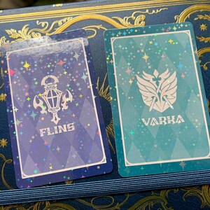 May include: Two trading cards with a holographic effect. One card is blue with a white lantern design and the word "FLINS". The other card is teal with a white emblem and the word "VARKA". Both cards have a white border and star accents.