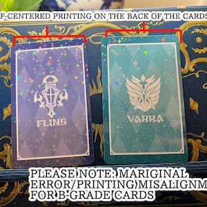 May include: Two playing cards with a holographic design. One card is blue with the word "FLINS" and a lantern illustration. The other is teal with the word "VARKA" and a crest. The cards have printing misalignment.