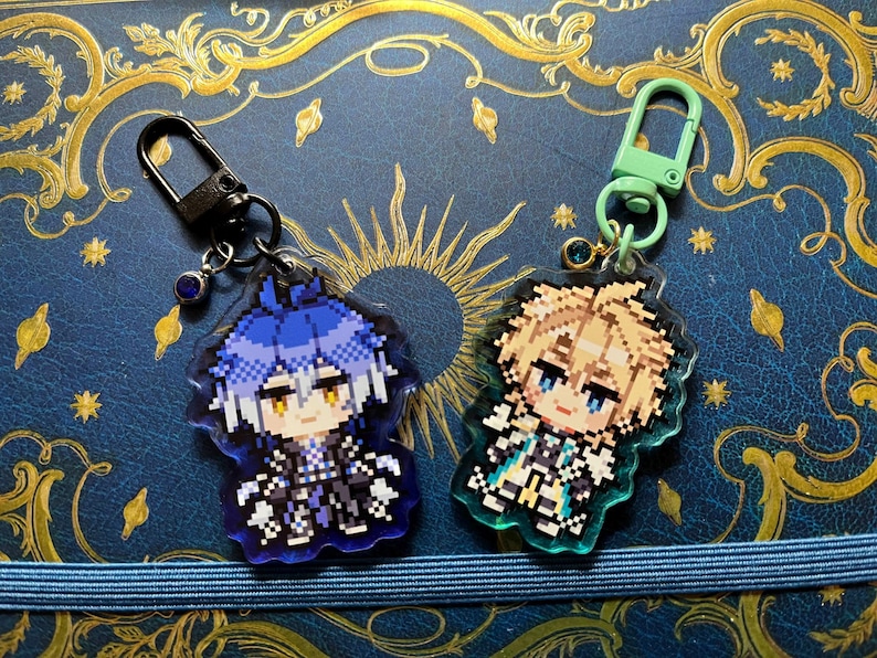 May include: Two pixel art character keychains. One keychain has a black clasp and a blue-haired character. The other keychain has a green clasp and a blonde-haired character. Both keychains are on a blue background with gold accents.