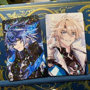 May include: Two rectangular anime-style character cards. One features a blue-haired character with a black and blue outfit, the other a blonde-haired character in a white and black outfit. Both cards have a holographic, sparkling effect.