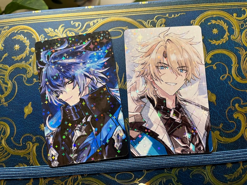 May include: Two rectangular anime-style character cards. One features a blue-haired character with a black and blue outfit, the other a blonde-haired character in a white and black outfit. Both cards have a holographic, sparkling effect.