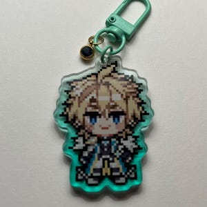 May include: A pixelated anime-style character keychain. The character has blonde hair and blue eyes, with a teal border. The keychain includes a teal clasp and a small gold charm with a dark blue gem. The overall design is colorful and detailed.