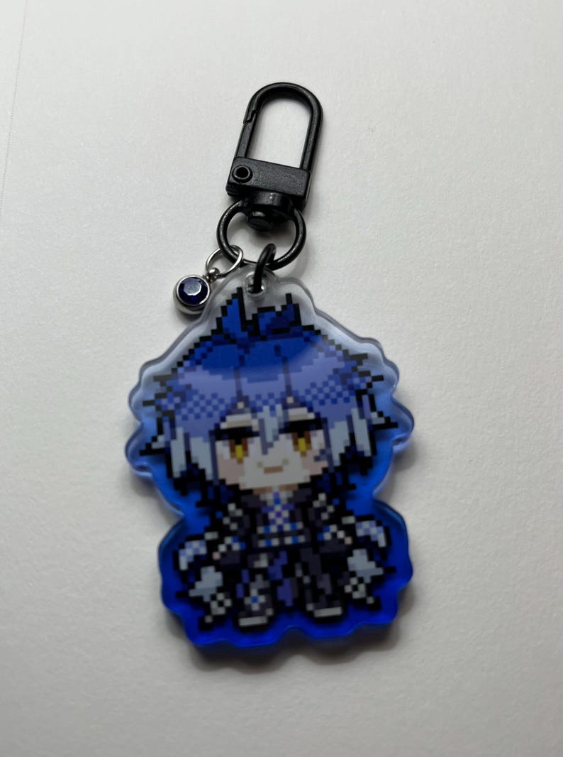 May include: A pixelated keychain featuring a character with blue hair and a black outfit. The keychain has a black clasp and a small charm. The character is set against a blue background.