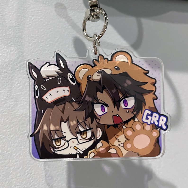 May include: A rectangular acrylic keychain featuring a cartoon illustration of four characters. One character is in a bear costume, another is a black horse-like creature, and two others are human-like figures. The word "GRR" is printed on the keychain.