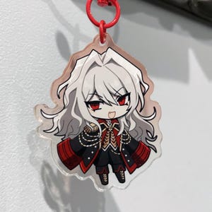 May include: A clear acrylic charm featuring a chibi anime character with white hair and red eyes. The character wears a black and red military-style outfit with gold accents. The charm hangs from a red metal ring.