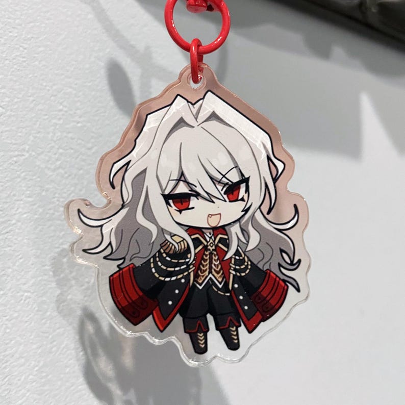 May include: A clear acrylic charm featuring a chibi anime character with white hair and red eyes. The character wears a black and red military-style outfit with gold accents. The charm hangs from a red metal ring.