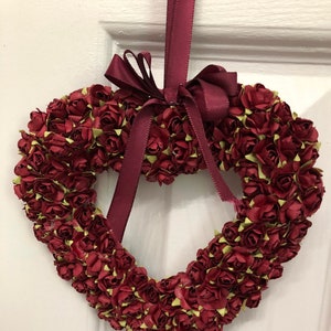 May include: A heart-shaped wreath made of burgundy red paper roses with green centers. The wreath is tied with a burgundy red satin ribbon bow.