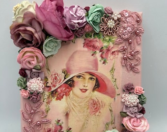 May include: A pink canvas featuring a vintage image of a woman in a hat, surrounded by pink, green and white flowers. The canvas is adorned with lace and pearls.