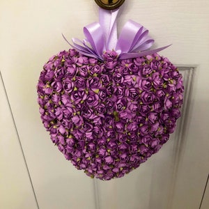 May include: A purple heart-shaped wreath made of paper roses with a lavender ribbon bow.