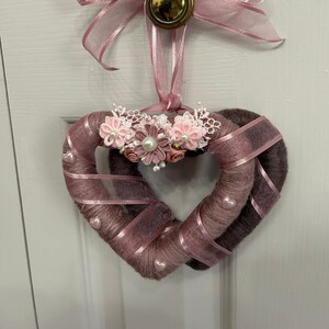 May include: A pink and brown yarn heart wreath with pink ribbon and lace flowers. The wreath is hanging from a gold knob on a white door.