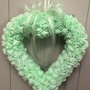May include: A heart-shaped wreath made of light green artificial roses with a white satin ribbon bow.
