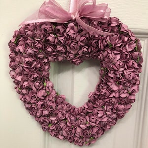 May include: A heart-shaped wreath made of small, pale pink paper roses. The wreath is tied with a pink ribbon and bow.