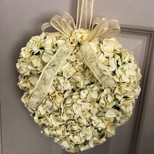 May include: A heart-shaped wreath made of white paper flowers with a gold ribbon and bow.