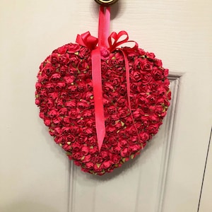 May include: A heart-shaped wreath made of pink paper roses with a pink ribbon hanging from a gold doorknob.