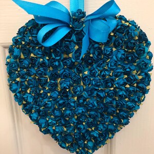 May include: A teal blue heart-shaped wreath made of small paper roses. The wreath has a teal blue ribbon tied in a bow at the top.