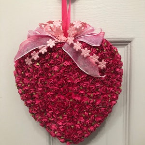 May include: A pink heart-shaped wreath made of small pink paper roses with a pink ribbon and bow.
