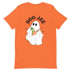 以下が含まれることがあります： オレンジ色のTシャツに、緑色の飲み物を手にした白い幽霊と、黒い文字で「BOO-JEE」と書かれている