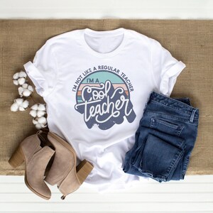 May include: White t-shirt with a retro-style graphic that says "I'm not like a regular teacher, I'm a cool teacher". The graphic is in a circle with a blue, green, and pink color scheme.
