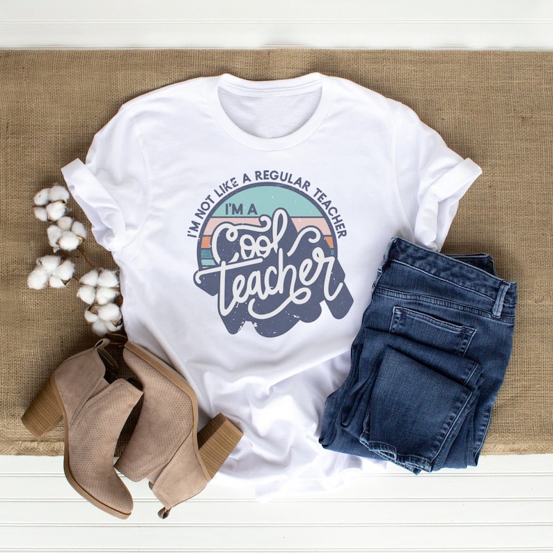 May include: White t-shirt with a retro-style graphic that says "I'm not like a regular teacher, I'm a cool teacher". The graphic is in a circle with a blue, green, and pink color scheme.