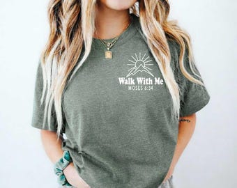 Könnte beinhalten: Ein olivgrünes, meliertes Kurzarm-T-Shirt mit einer weißen Grafik auf der linken Brust. Das Design zeigt einen Sonnenaufgang über einem Bergumriss mit dem Text "Walk With Me" und "MOSES 6:34" in einer klaren, weißen Schriftart.