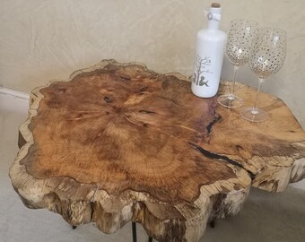 May include: A rustic wooden coffee table with a live edge top and black metal legs. The table is made from a tree trunk slice and has a natural wood grain pattern. Two clear glass wine glasses with gold polka dots are on the table, along with a white bottle with a decorative design.