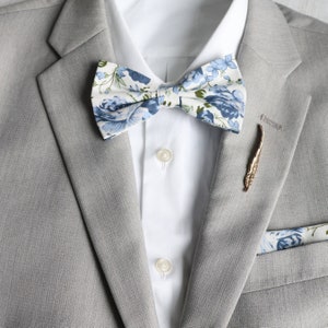 Peut inclure: Une veste de costume grise avec une chemise blanche, un nœud papillon floral bleu et blanc et une épingle en or en forme de plume.