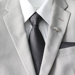 May include: A gray suit jacket with a black patterned tie and a silver pin. The jacket has a pocket square with a matching pattern.