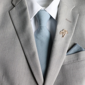 May include: A gray suit jacket with a light blue tie and a gold pin with a tree design. The suit jacket has a pocket square in the breast pocket.