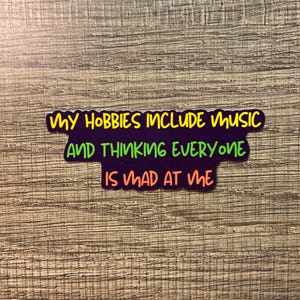 May include: A sticker with the text "MY HOBBIES INCLUDE MUSIC AND THINKING EVERYONE IS MAD AT ME" in yellow and green on a purple background. The sticker is on a wooden surface.