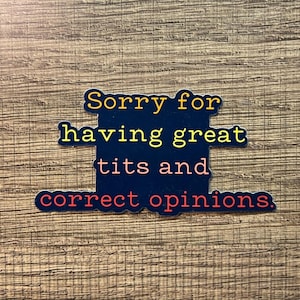 May include: A navy blue sticker with the text "Sorry for having great tits and correct opinions" in yellow and red lettering. The sticker is rectangular with rounded corners and is set against a wood grain background.