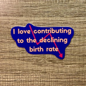 May include: A blue sticker with the text "I love contributing to the declining birth rate" in orange. A red line graph points downward, emphasizing the decline. The sticker is on a wooden surface.