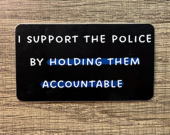 May include: A black rectangular card with rounded corners. The white text reads "I SUPPORT THE POLICE BY HOLDING THEM ACCOUNTABLE." The words "HOLDING THEM" are highlighted in blue. The card is on a wooden surface.