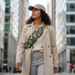 Könnte beinhalten: Eine Frau trägt einen beigen Trenchcoat, eine hellgraue Jeans und eine grüne Bauchtasche mit floralem Muster. Sie trägt auch eine beige Baseballkappe. Der Hintergrund zeigt eine Stadtstraße mit hohen Gebäuden.
