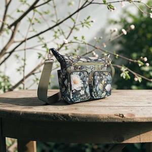 Könnte beinhalten: Eine Umhängetasche mit floralem Muster, einem verstellbaren grünen Riemen und goldfarbener Hardware. Die Tasche hat zwei Vordertaschen mit Reißverschlüssen und ein Hauptfach. Das florale Muster umfasst weiße Blumen und grüne Blätter. Die Tasche liegt auf einem Holztisch.