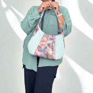 Könnte beinhalten: Schultertasche mit floralem Muster und hellblauen, strukturierten Akzenten. Die Tasche hat einen Reißverschluss und hellblaue Träger. Das Blumenmuster weist rosa, pfirsichfarbene und blaue Farbtöne auf. Die Person trägt ein hellgrünes Hemd und eine dunkelblaue Hose.