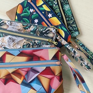 Könnte beinhalten: Vier verschiedene Stoff-Reißverschlusstaschen mit braunen Lederelementen. Die Taschen haben verschiedene Muster, darunter tropische Blätter, geometrische Formen und Früchte. Jede Tasche hat zwei Reißverschlüsse und einen Handgelenksriemen.