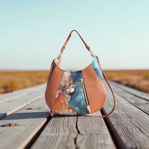 Könnte beinhalten: Eine braune Hobo-Tasche aus Leder mit einem farbenfrohen, abstrakten Design. Die Tasche hat eine Reißverschlusstasche und einen langen Riemen. Das Design enthält Farbtöne von Blau, Orange und Gold. Die Tasche liegt auf einer Holzoberfläche.
