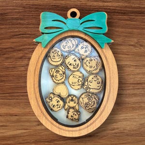 May include: Wooden oval ornament with a teal bow and cartoon faces inside. The light-coloured wood ornament has a small hole for hanging. The faces are etched into the wood, creating a decorative and unique design.